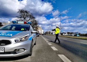 Zdjęcie poglądowe do komunikatu, które przedstawia umundurowanego policjanta ruchu drogowego, zatrzymującego samochód. W kadrze widać policyjny, oznakowany radiowóz.