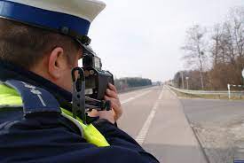 Zdjęcie poglądowe przedstawiające umundurowanego policjanta ruchu drogowego dokonującego pomiaru prędkości ręcznym miernikiem prędkości. na zdjęciu widać skrzyżowanie dróg. Jest to zdjęcie poglądowe. Zdjęcie pobrano ze strony www.kolobrzeg.policja.gov.pl
