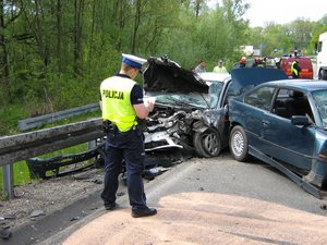 Zdjęcie poglądowe przedstawia policjanta ruchu drogowego wykonującego czynności służbowe na miejscu zdarzenia. na zdjęciu widoczny jest również rozbity samochód