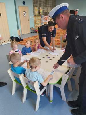 Na zdjęciu widać umundurowanych policjantów podczas zajęć z dziećmi w sali przedszkolnej. Dzieci rysują a policjanci częstują je słodyczami.