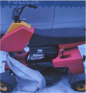 zdjęcie przedstawia kosiarkę samojezdną w kolorze czerwono żółtym w przedniej części kosiarki widoczna plandeka kol. siwego. Kosiarka stoi na tle oznakowanego policyjnego radiowozu.