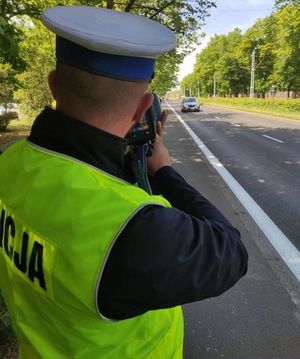 Zdjęcie poglądowe przedstawiające umundurowanego funkcjonariusza ruchu drogowego stojącego przy drodze i dokonującego pomiaru prędkości pojazdów przy użyciu ręcznego miernika prędkości