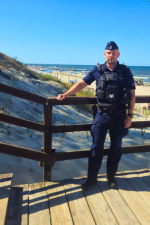 Zdjęcie przedstawia umundurowanego Policjanta SPPP w Częstochowie stojącego na schodach nadmorskiego zejścia w tle widać plażę i morze. Policjant dwukrotnie uratował życie chłopca i dziewczynki.