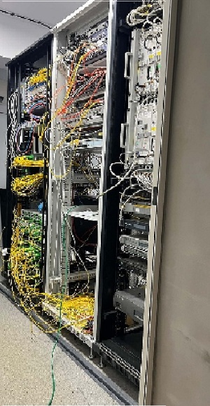Zdjęcie przedstawia pomieszczenie z metalowymi regałami a na nim stojące urządzenia i okablowanie służące do rozpowszechniania sygnału